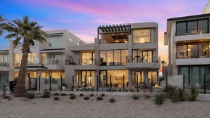 A modern multi-story beachfront property with large windows, outdoor seating, and a well-maintained garden.