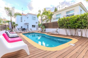 Una piscina con sillas de descanso, una terraza de madera y un edificio blanco con balcones.