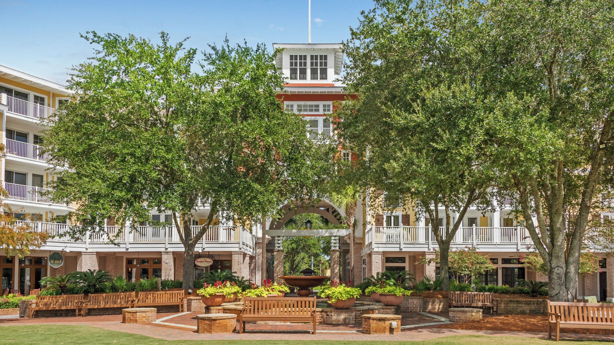 A courtyard with a central fountain, surrounded by benches and potted plants, with buildings in the background.