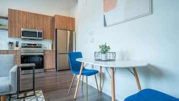 A modern kitchen with wooden cabinets, a stainless steel refrigerator, and a microwave. There is a dining area with a white table and blue chairs.