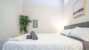 En snyggt bäddad säng med ett vitt, vadderat täcke, en grå filt och två hoprullade handdukar. På sängens vänstra sida står en liten växt, och på väggen sitter en strömbrytare.