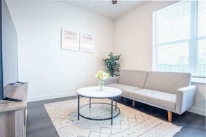 A living room with a sofa, a round coffee table, a TV on a stand, a plant, and a window with blinds. - Boulevard Stays Comfortable 1BR Suite w Pool, Parking, Gym 05 (Dallas)