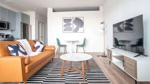 A modern living room with a sofa, a round table, two chairs, a TV, and a kitchen area in the background. - Comfortable 1BR Suite w Pool, Parking, Gym 08 (Dallas)
