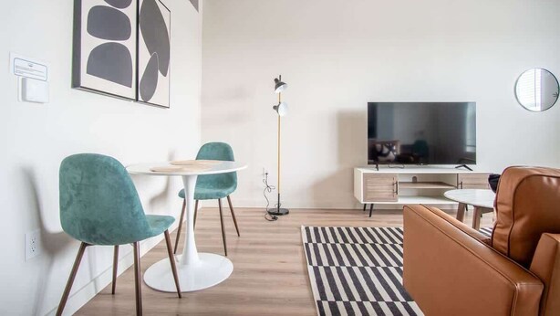 A modern living room with a flat-screen TV, teal chairs, a white coffee table, a brown leather sofa, and a black and white striped rug. - Comfortable 1BR Suite w Pool, Parking, Gym 08 (Dallas)