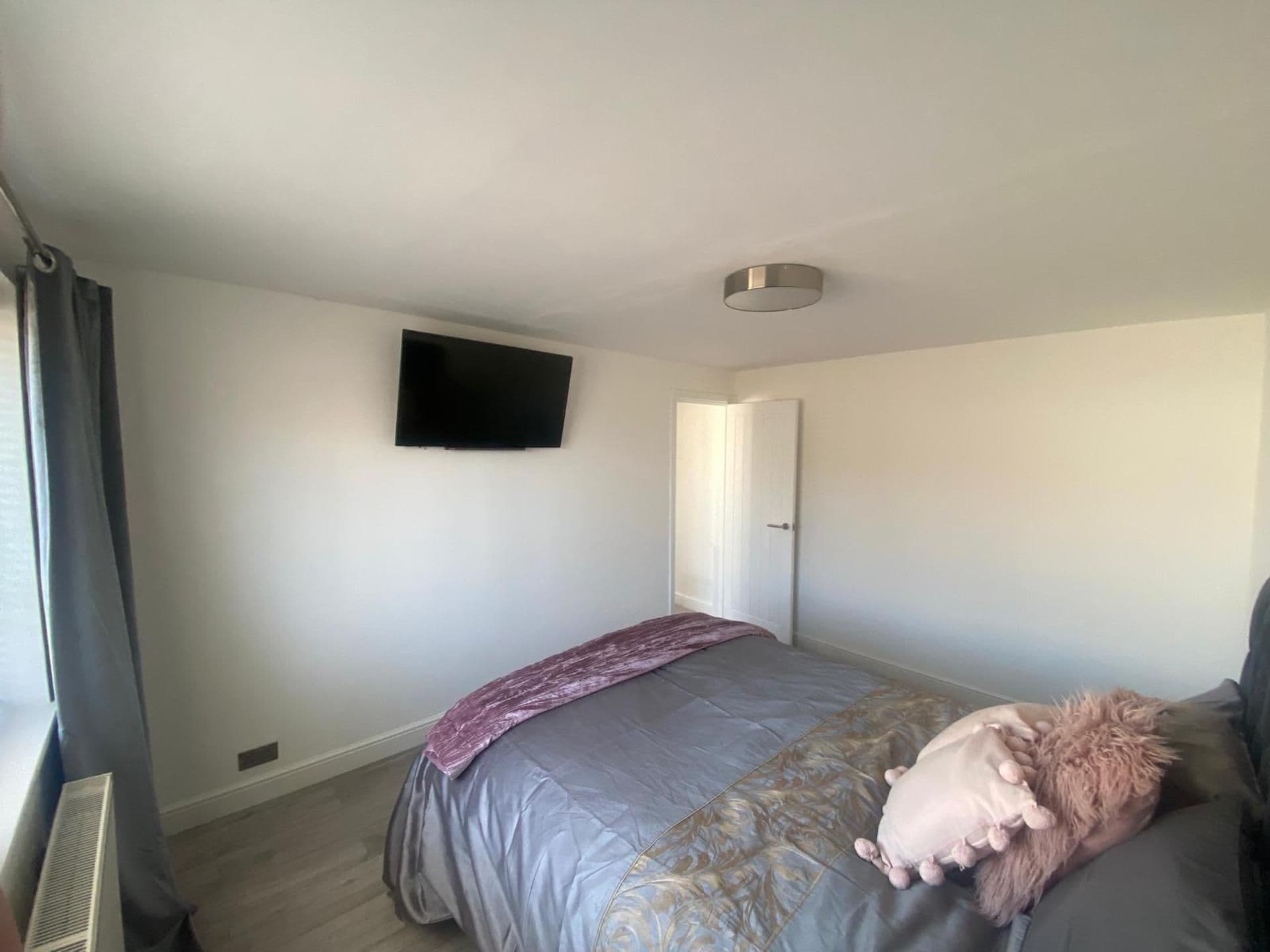 A bedroom with a bed, a flat-screen TV mounted on the wall, and a door leading to another room.
