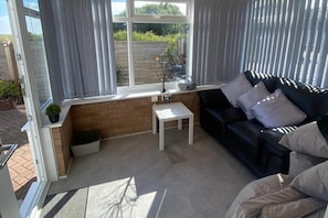 A sunlit room with a black sofa, a small white table, and a window with curtains. - Prestatyn 2-bed Bungalow, Sleeps 5 With Hot Tub (Prestatyn)