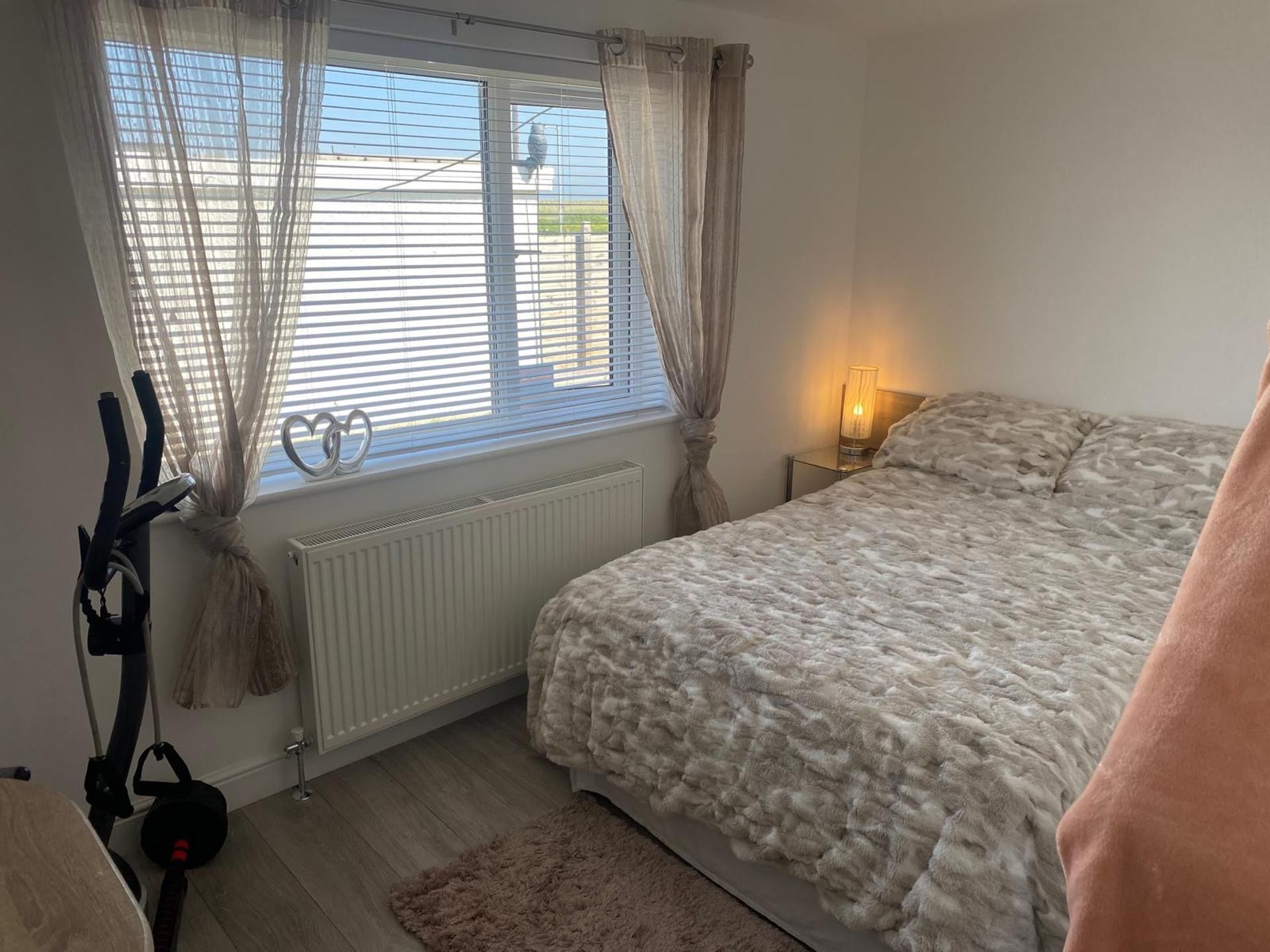 A bedroom with a bed, a radiator, a window with curtains, a nightstand with a lamp, and exercise equipment.