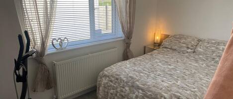 A bedroom with a bed, a radiator, a window with curtains, a nightstand with a lamp, and exercise equipment.