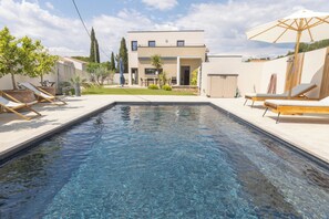 Une piscine entourée de chaises longues et de parasols, le tout bordé d’une maison moderne dotée d’un jardin.