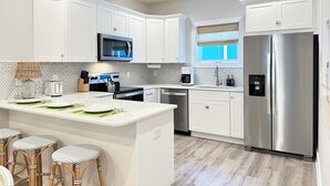 Una cocina moderna con armarios blancos, electrodomésticos de acero inoxidable, una isla central con taburetes y un ventanal con persianas.