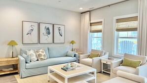 Un salón con un sofá azul claro, una mesa de centro blanca y dos sillones. En la pared hay cuadros enmarcados de pájaros y una ventana con cortinas.