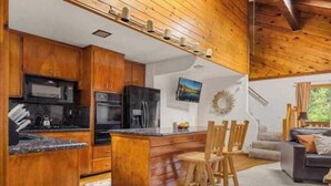 Una cocina con armarios de madera, un refrigerador negro y un microondas. Hay un comedor con sillas y una mesa de madera.