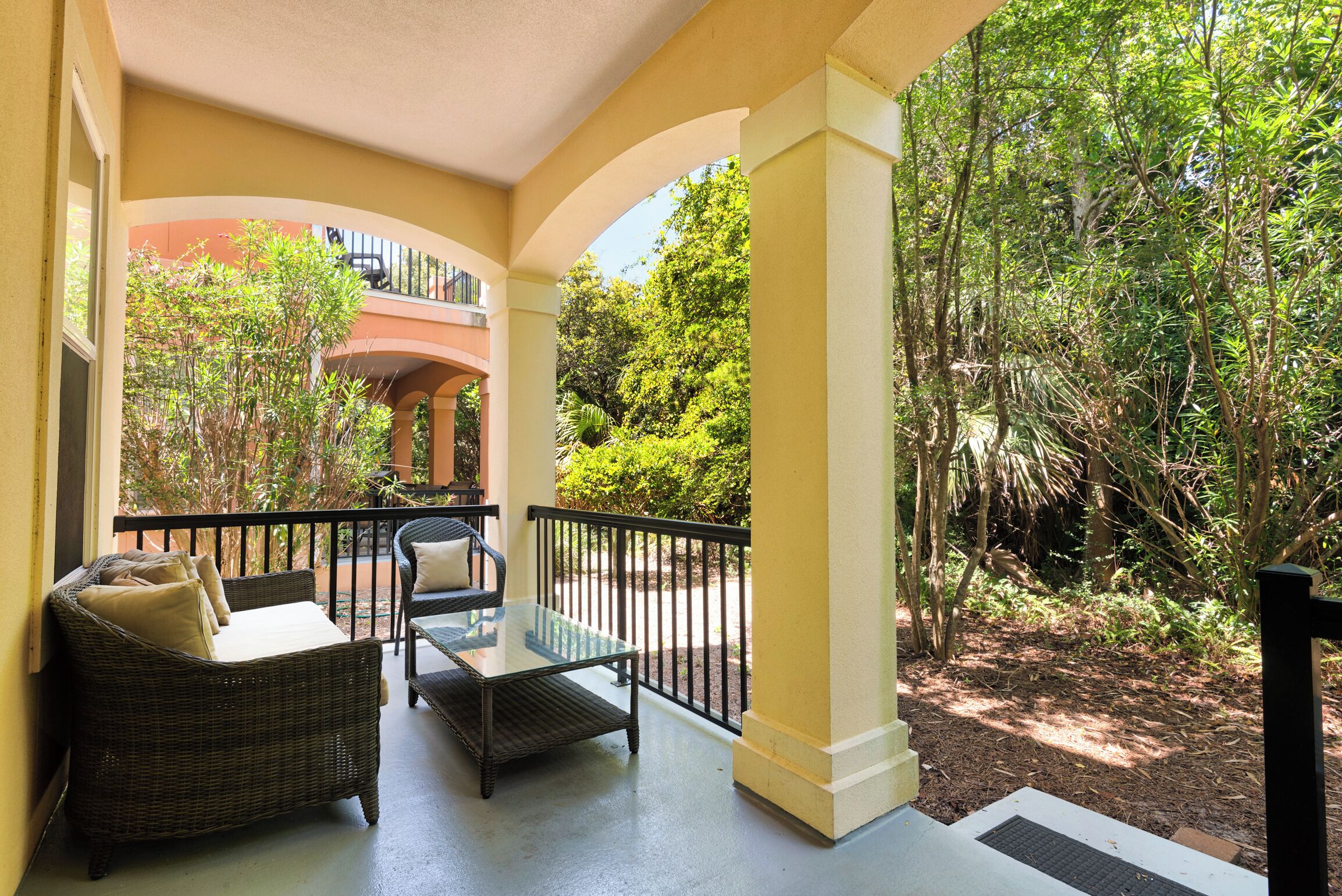 A covered outdoor seating area with wicker furniture and a glass table, surrounded by lush greenery and a building in the background.