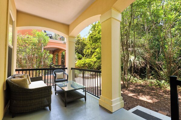 A covered outdoor seating area with wicker furniture and a glass table, surrounded by lush greenery and a building in the background.