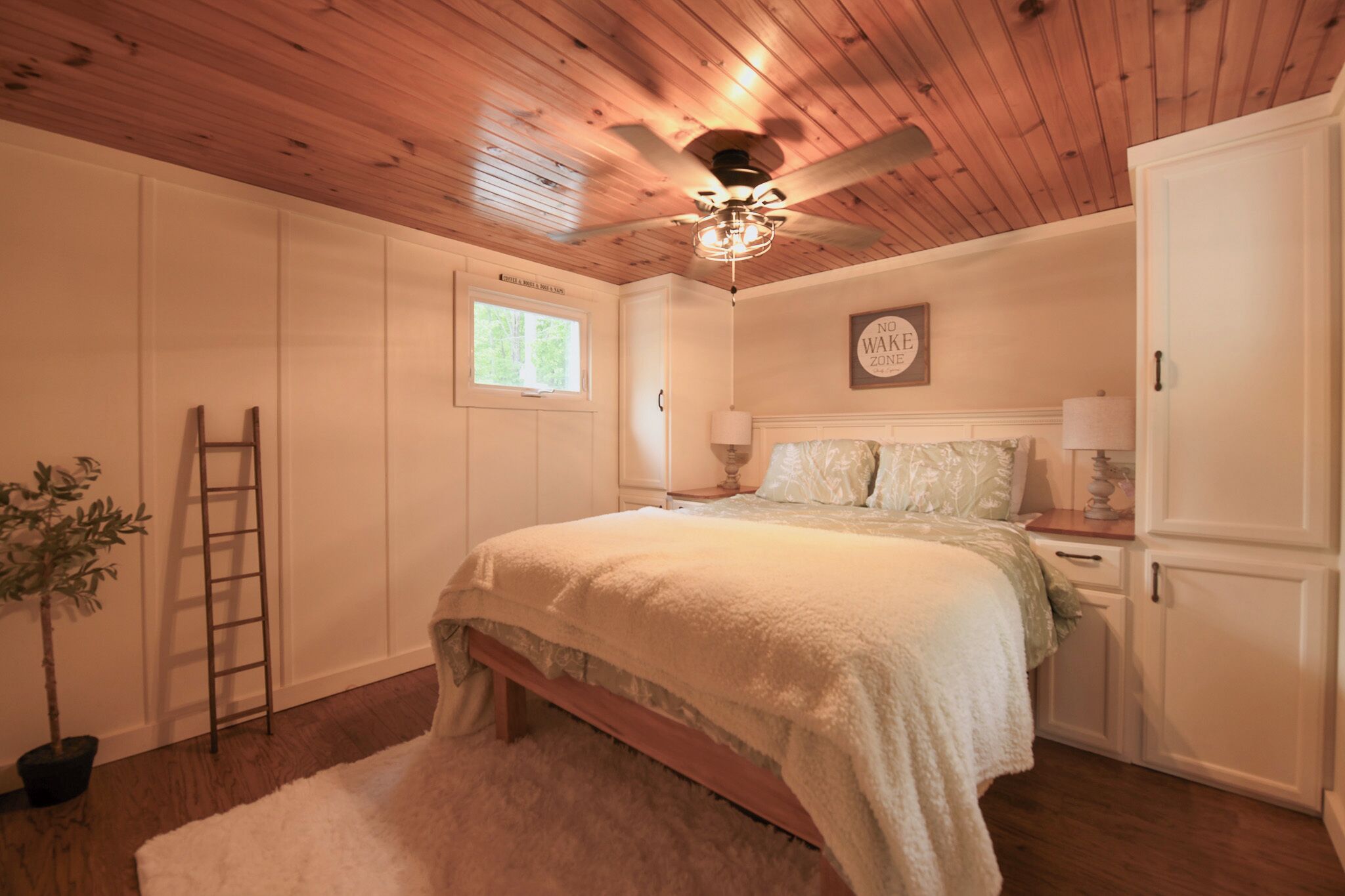Una camera da letto con soffitto in legno, una ventola a soffitto, un letto con copripiumino bianco, un comodino, una lampada, una cassettiera, una scala e una pianta in vaso.