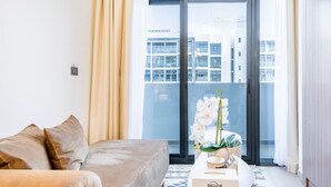 En moderne stue med en sofa, et sofabord med en æske med teksten "DELUXE" og udsigt til en bygning gennem et stort vindue.