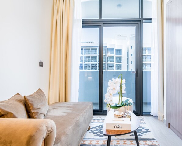 A modern living room with a sofa, a coffee table with a box labeled "DELUXE," and a view of a building through a large window. - Charming Studio at Azizi Riviera 32, MBR City by Deluxe Holiday Homes (Dubai)