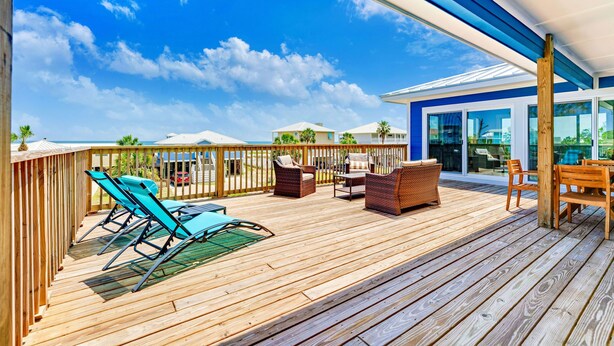 Een houten terras met tuinmeubilair, waaronder stoelen en een tafel, met uitzicht op een helderblauwe lucht en een woonwijk met huizen.