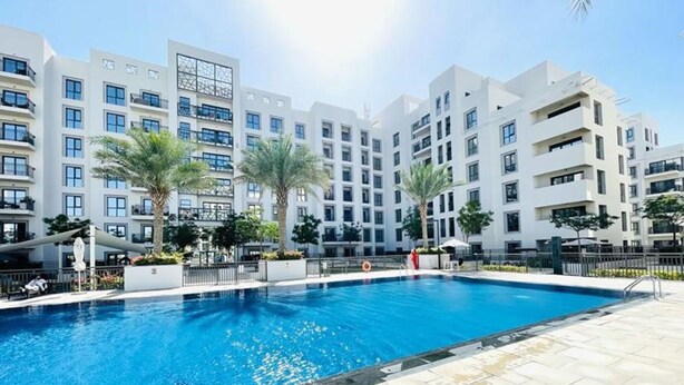 A swimming pool surrounded by modern buildings with balconies, palm trees, and a clear sky.
