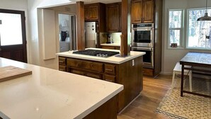 A modern kitchen with wooden cabinets, a central island, stainless steel appliances, and a view of the outdoors through large windows.