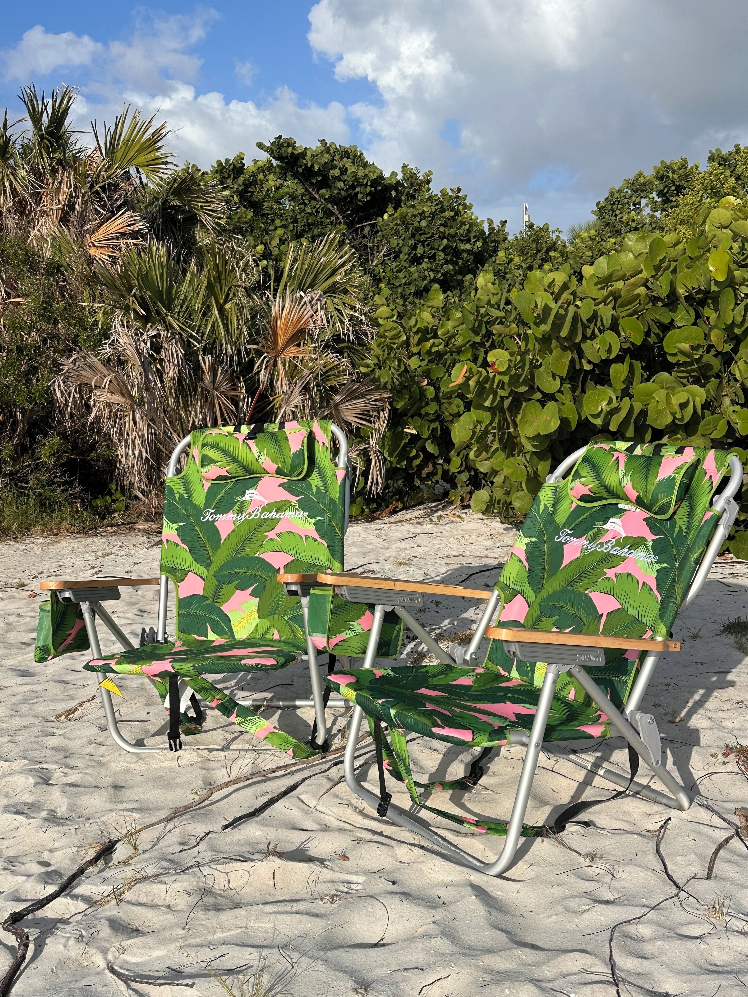 Two beach chairs with a tropical leaf pattern, positioned on a sandy beach with dense foliage in the background.