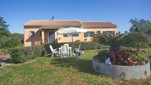 A two-story house with a tiled roof, a front porch with chairs and an umbrella, and a garden with plants and a palm tree. - Eleni's House by Corfuescapes (Corfu)