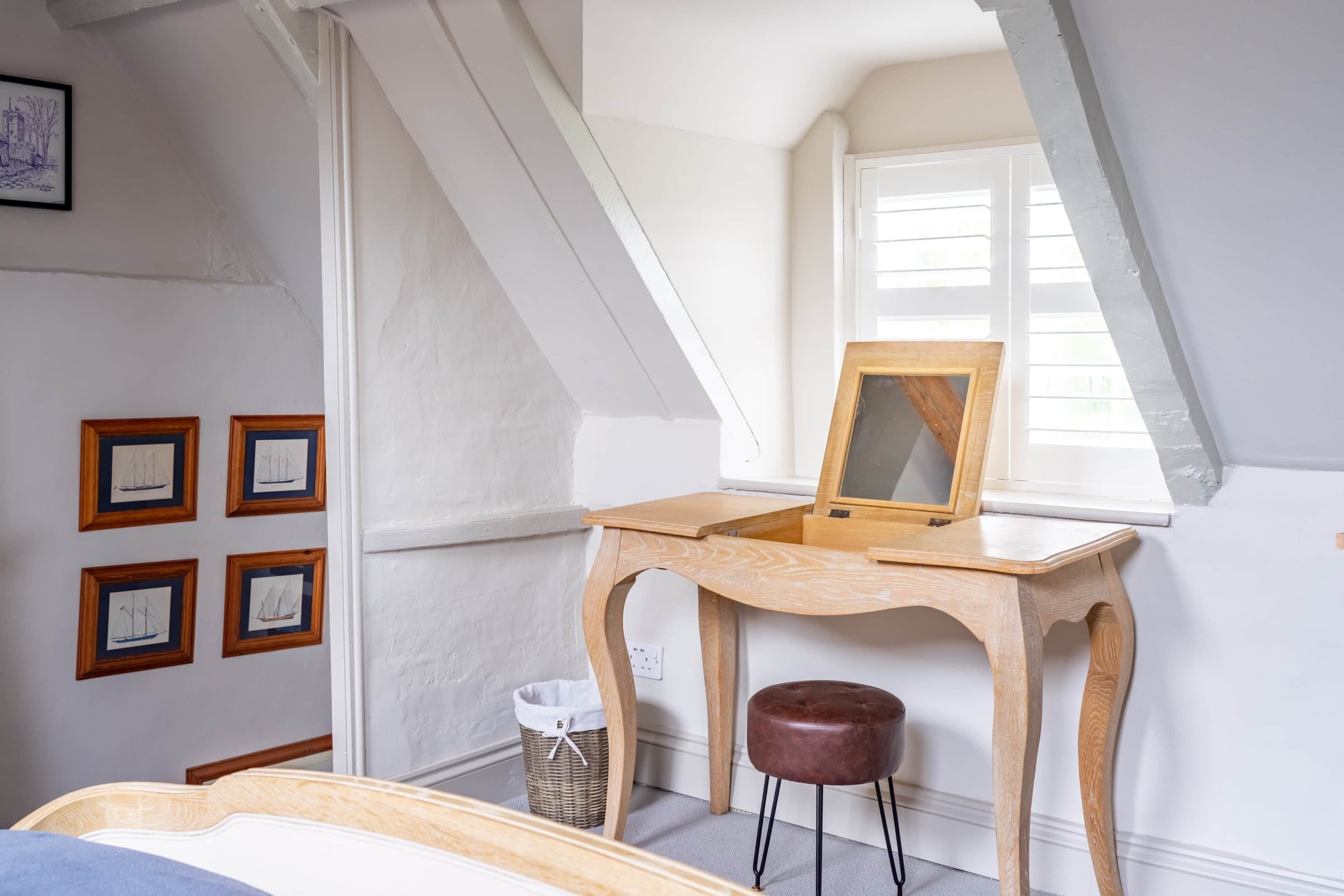 A bedroom with a sloped ceiling, a wooden desk with a mirror, a wicker basket, and framed pictures on the wall.