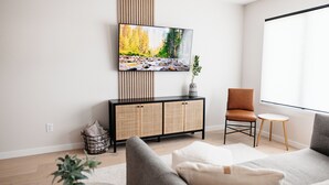 A modern living room with a flat-screen TV mounted above a wicker console, a grey sofa, a brown leather chair, and a small round table. - -The Cotton House- Modern, Clean, Close to MSU! (Mankato)