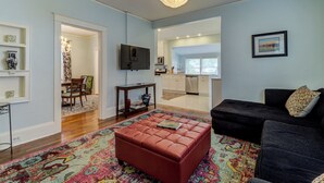 Ein Wohnzimmer mit einem roten Pouf, einem schwarzen Sofa, einer Deckenleuchte und einer Küche im Hintergrund.