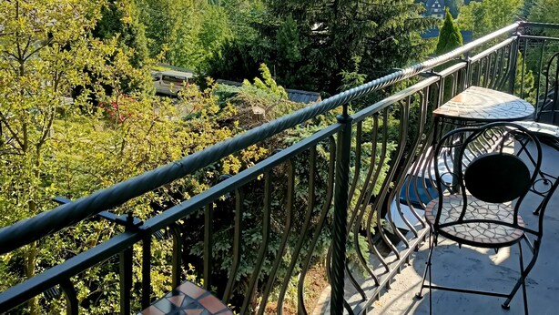 A balcony with a metal railing, a small table, and a chair, overlooking a lush green landscape with trees and distant mountains. - Cozy 3-Bedroom Apartment for 5-6 guests with mountain views, with free parking (Zakopane)