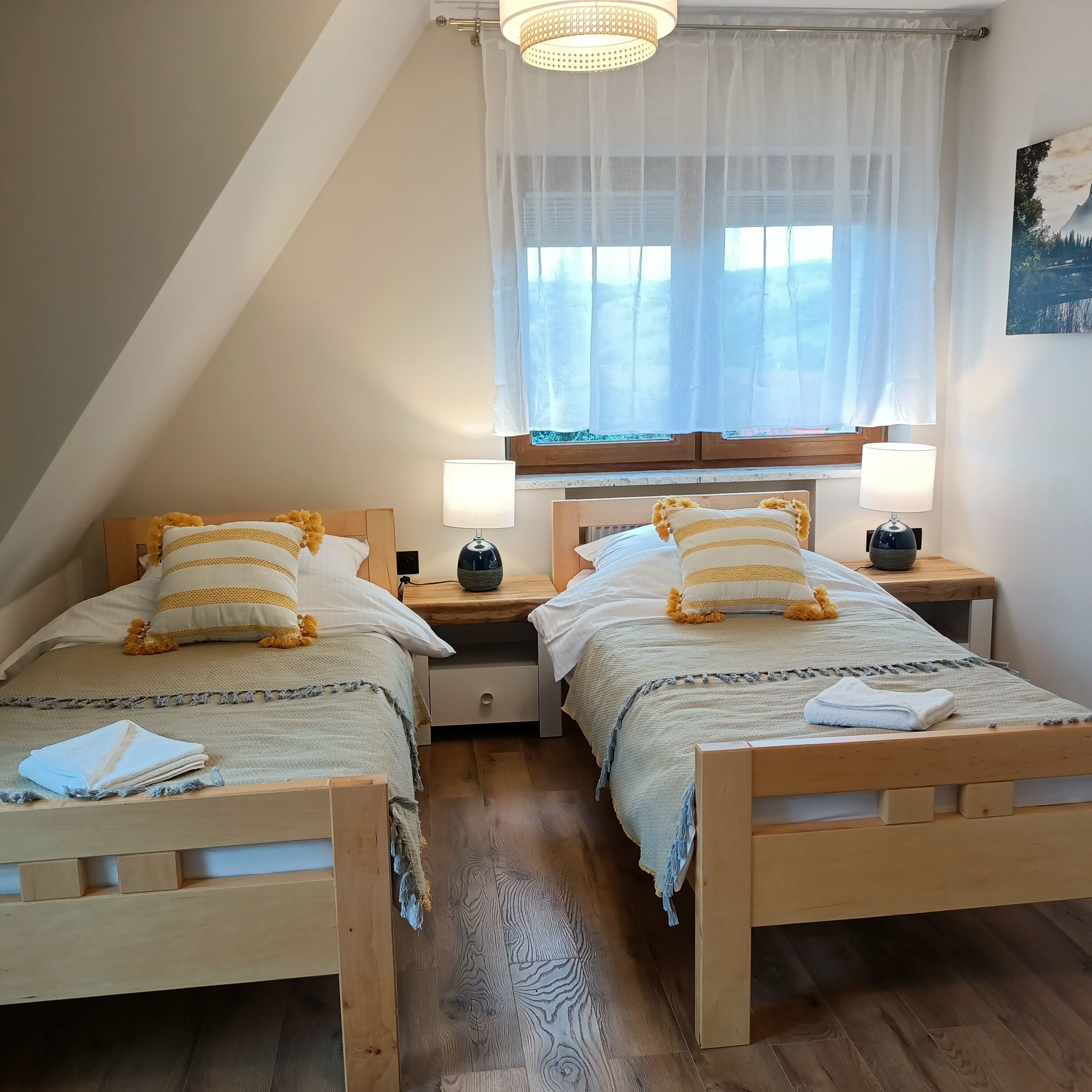 A bedroom with two wooden beds, each with a pillow and a bedside table with a lamp. There is a window with sheer curtains and a painting on the wall.