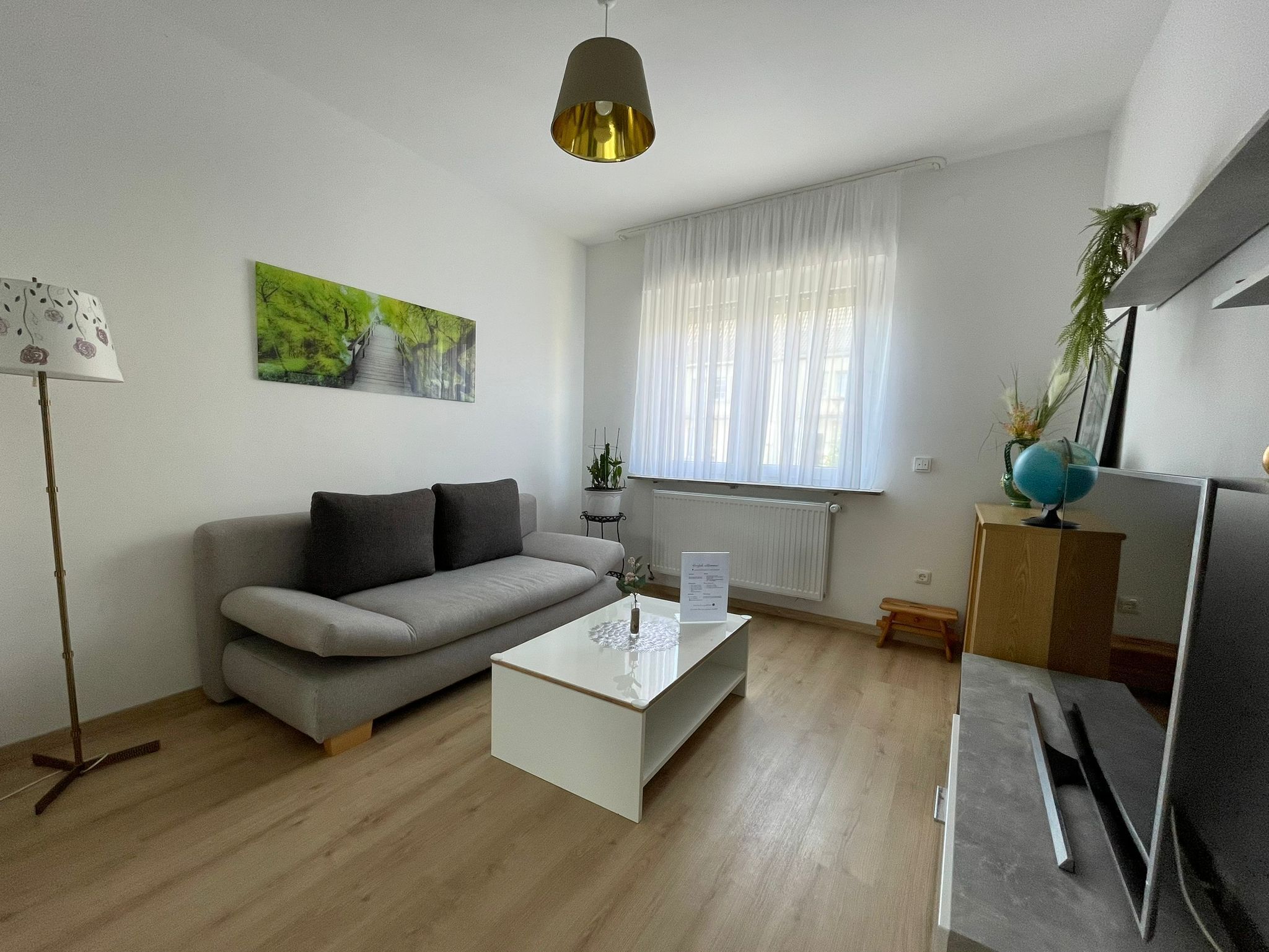Una sala de estar moderna con un sofá gris, una mesa de centro blanca, una lámpara de pie, un cuadro, una ventana con cortinas translúcidas, un radiador, un mueble de madera pequeño y un globo terráqueo en una estantería.
