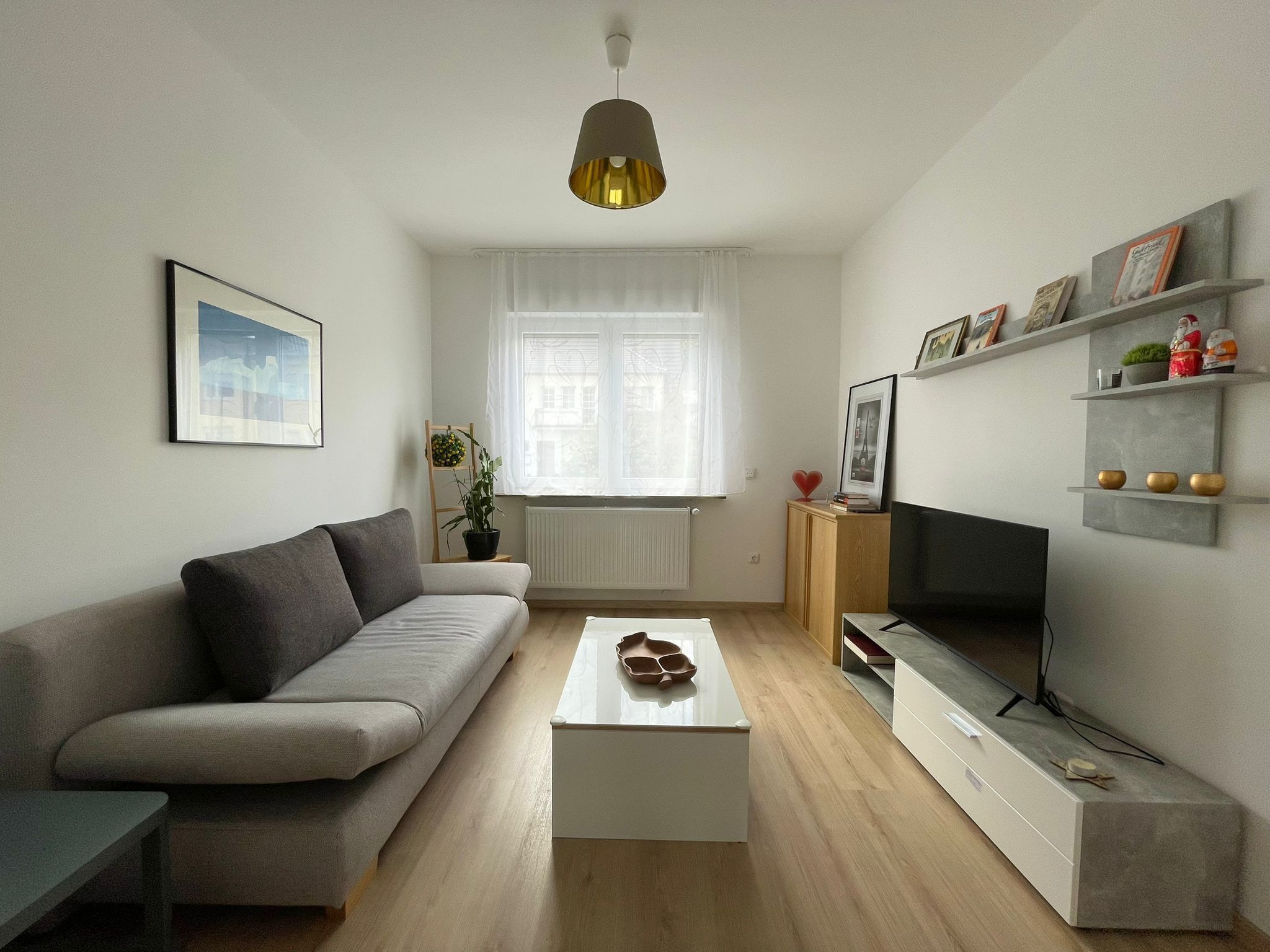Een moderne woonkamer met een grijze bank, een witte salontafel, een tv op een dressoir en een raam met dunne gordijnen.
