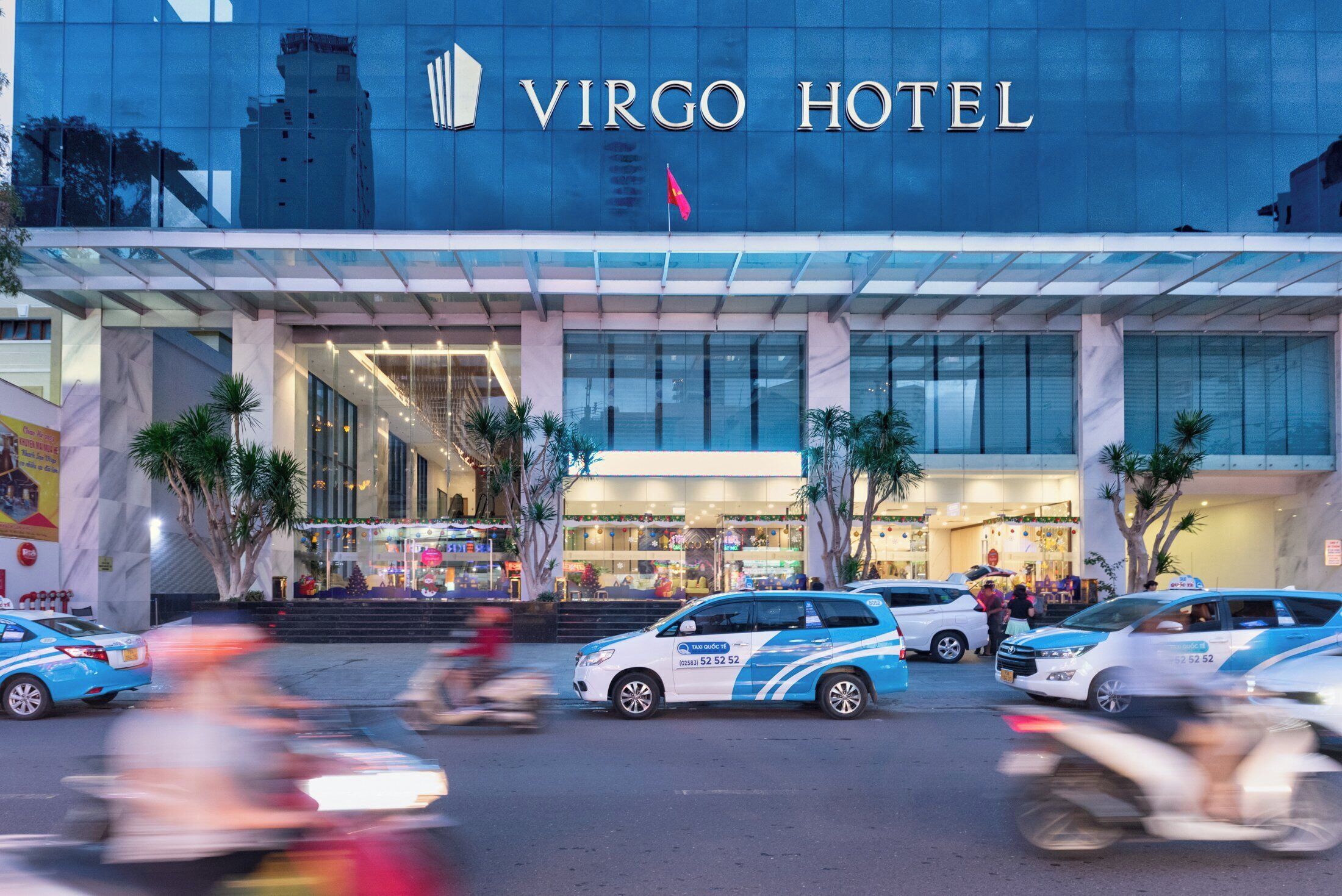 L’extérieur de l’hôtel Virgo se caractérise par sa façade moderne en verre, ses palmiers et une rue animée où circulent des voitures et des motos.
