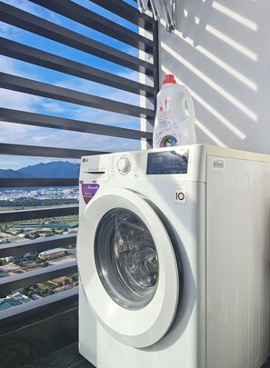 A front-loading washing machine with a detergent bottle on top, a digital display, and a view of a cityscape through a window with blinds. - S Lux Apartment Virgo Nha Trang (Nha Trang)
