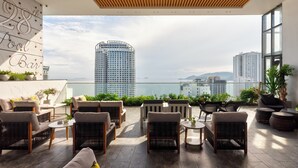 A rooftop lounge with wicker furniture, a pool bar sign, and a view of high-rise buildings and the sea. - S Lux Apartment Virgo Nha Trang (Nha Trang)