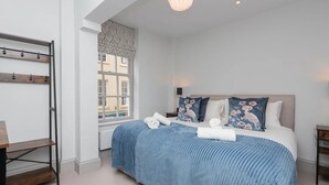 A bedroom with a bed, a wooden shelf, a window, and a hanging light fixture. - Stylish 2-bed apartment in the heart of Bath - Pass the Keys (Bath)