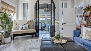 A spacious living room with a beige sofa, a blue sofa, and a black leather ottoman. There is a large window with a view of the outdoors, and a wooden ladder with decorative items on the right side. - Oak & Ocean * Pet Friendly, Boat Launch, Firepit, Hot Tub, Grill (Santa Rosa Beach)
