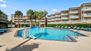 A swimming pool with lounge chairs, palm trees, and a multi-story hotel building in the background. - Salt Therapy, Great Amenities, Pool, Walk To Beach! (Galveston)