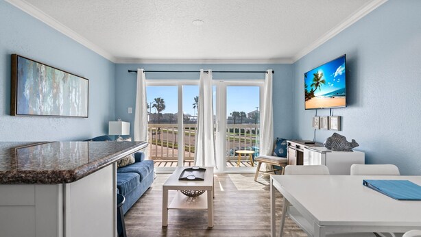 A modern living room with a large window, a flat-screen TV, and a dining area. - Salt Therapy, Great Amenities, Pool, Walk To Beach! (Galveston)