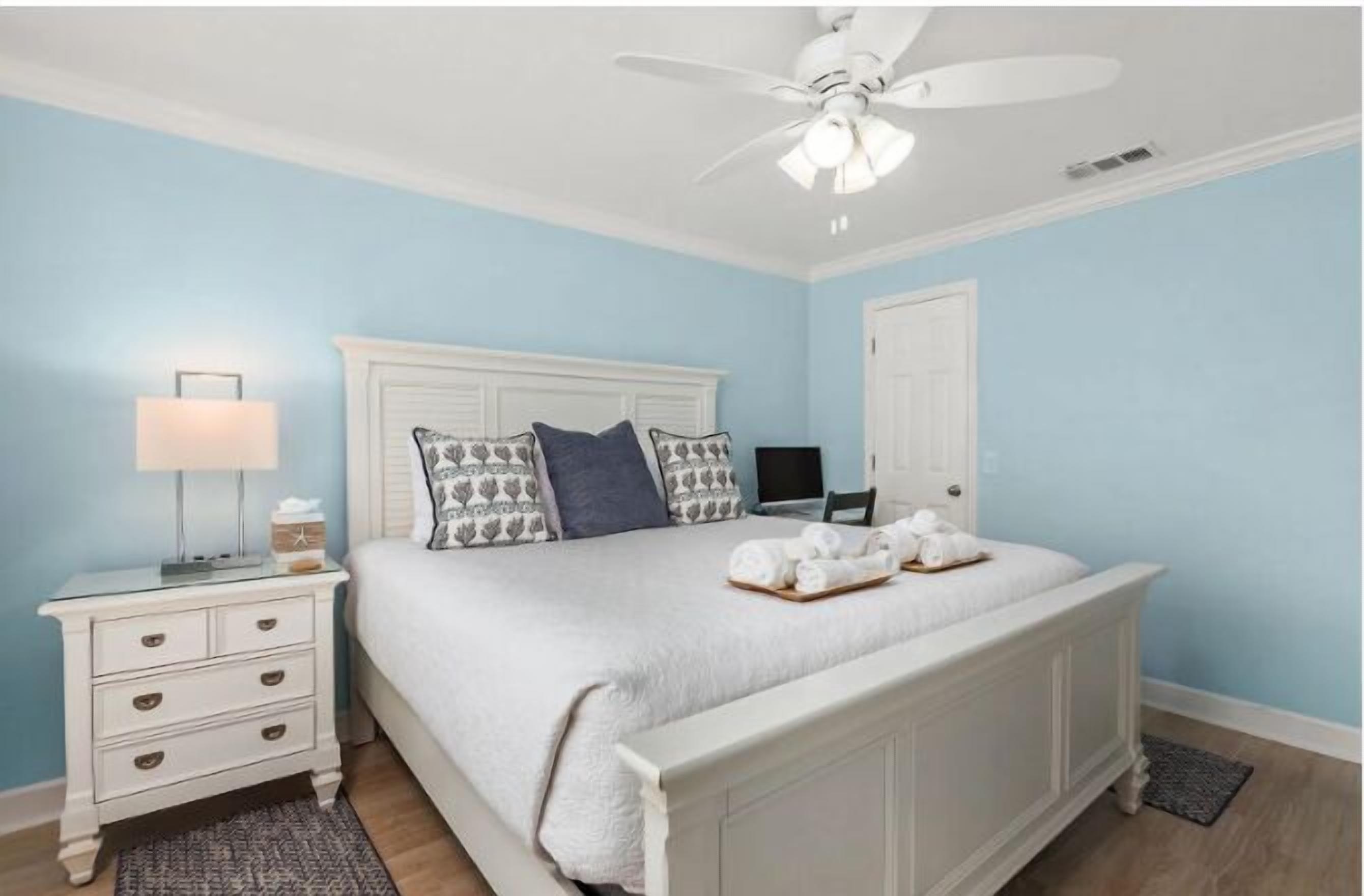 A bedroom with a white bed, a nightstand with a lamp, and a ceiling fan.