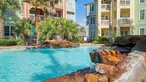 A swimming pool with a rock waterfall feature, surrounded by multi-story buildings with balconies and palm trees. - Ocean View Condo w/ resort amenities! (Galveston)