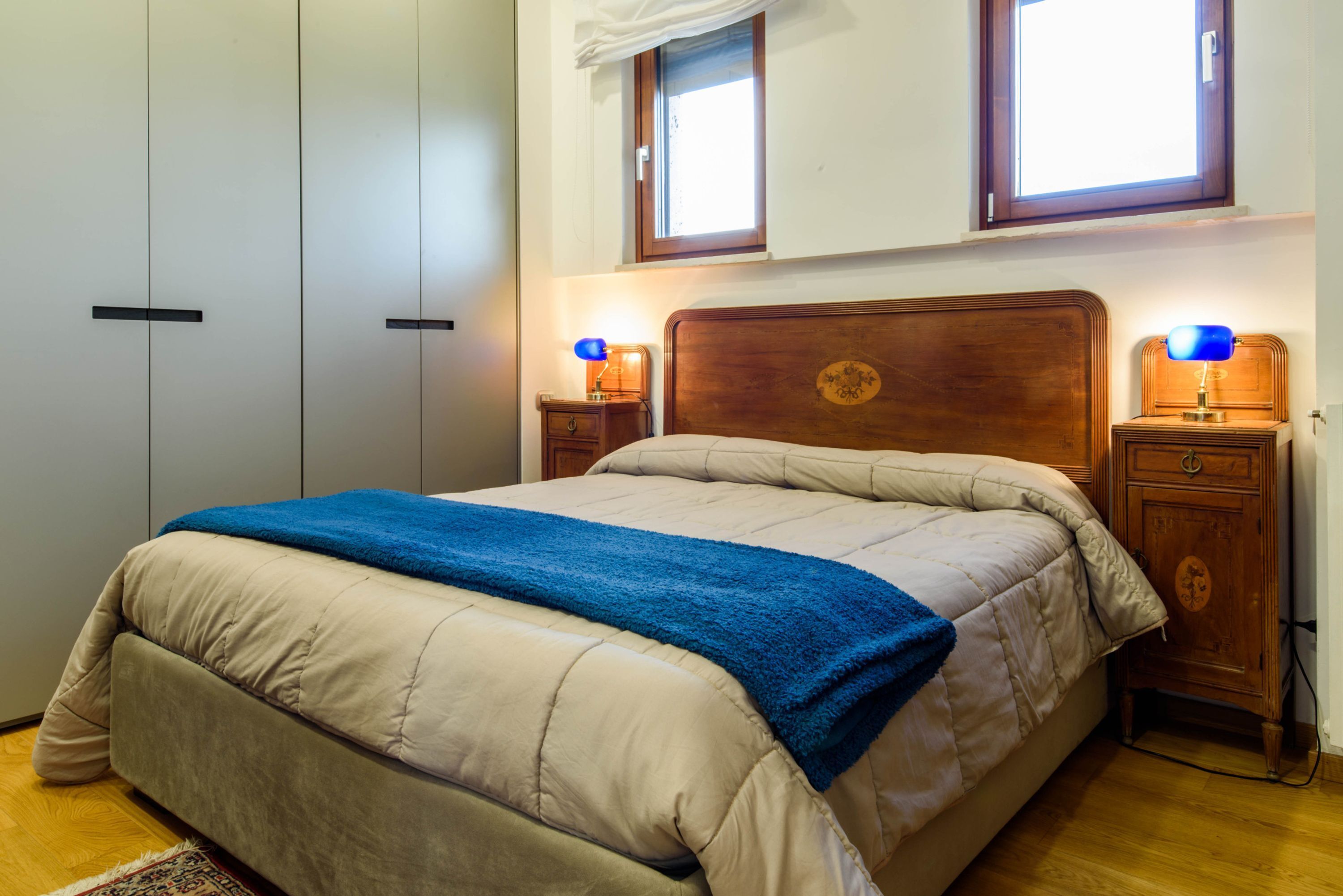 A bedroom with a bed, two bedside tables with lamps, a wardrobe, and a window with curtains.