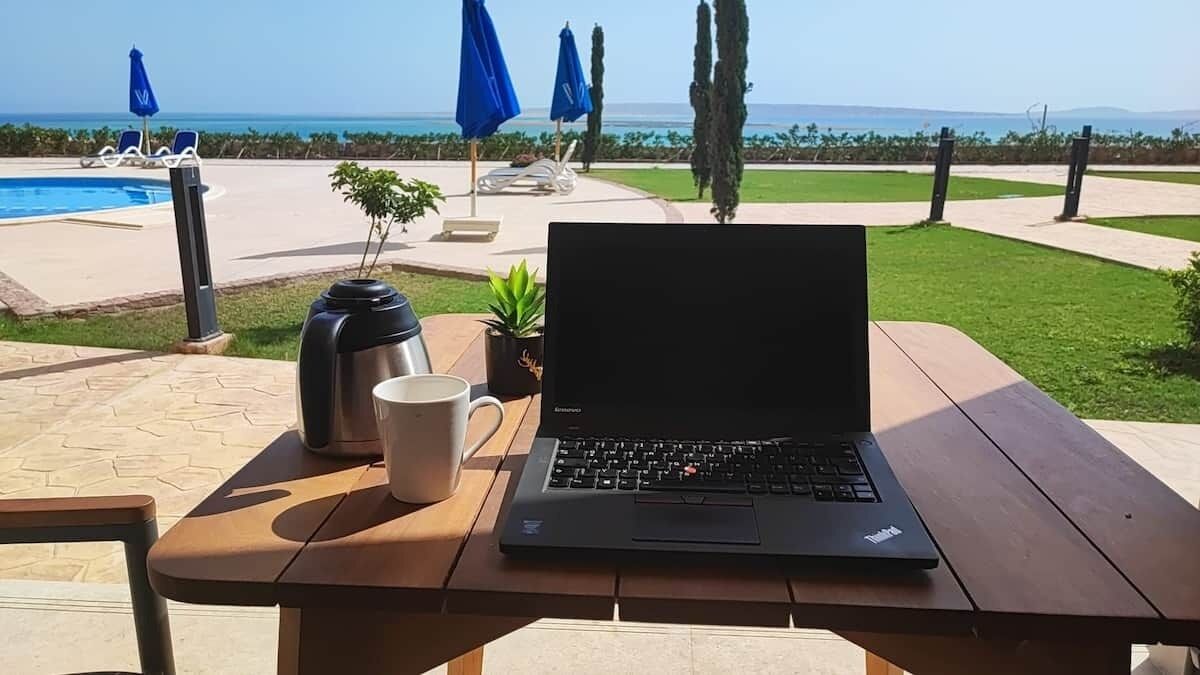 A laptop on a wooden table with a coffee mug and a thermos, overlooking a pool and greenery.