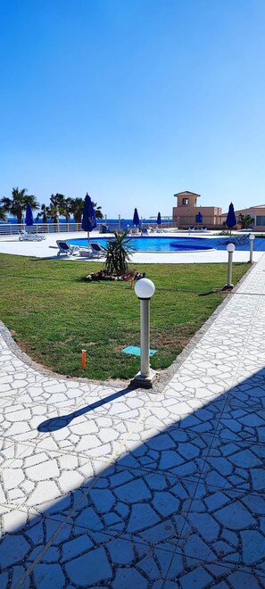 A pool area with lounge chairs and umbrellas, a clear sky, and a building in the background.