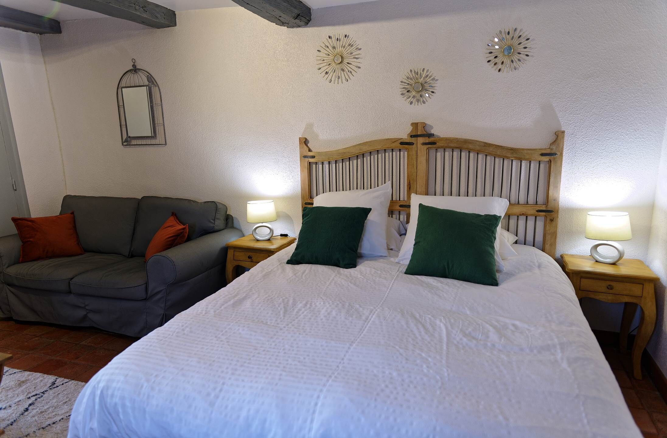 A bedroom with a large bed, a wooden headboard, two nightstands with lamps, a grey sofa with orange cushions, and decorative wall mirrors.