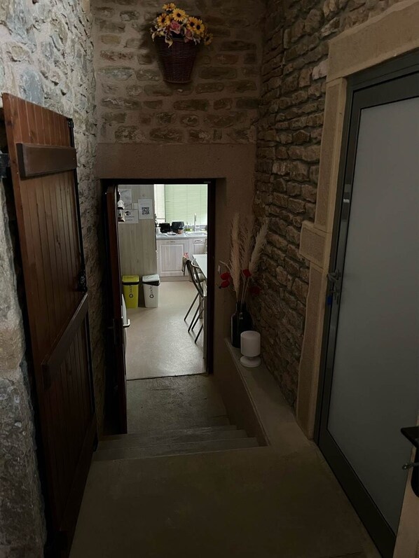 A narrow hallway with a stone wall and a wooden door on the left. A basket of flowers hangs on the wall, and a white door is on the right. - My little paradise / private indoor pool (Lournand)