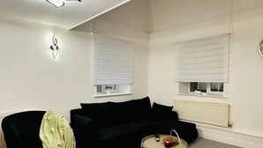 A modern living room with a black sofa, a round coffee table, and a black chair. There are two windows with blinds, a radiator, and a ceiling light fixture. - Home Away From Home- City Centre Gem (Derby)