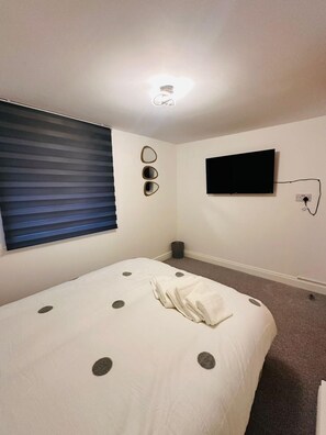 Ein Schlafzimmer mit einem Bett, einem Fernseher, einem Fenster mit Jalousien und einem an der Wand befestigten Spiegel.