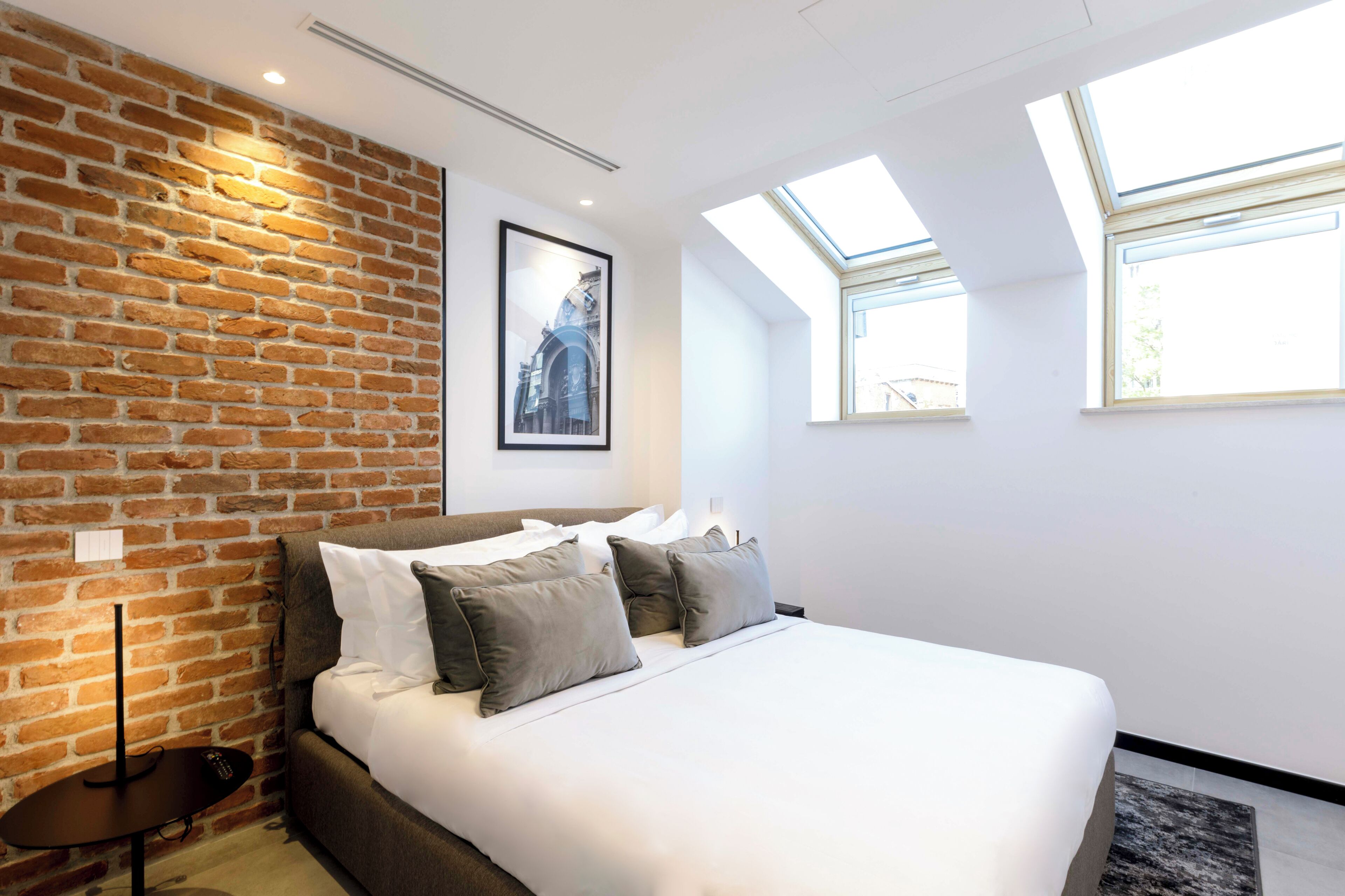 Un dormitorio con pared de ladrillo, cama con ropa de cama blanca y tragaluces.